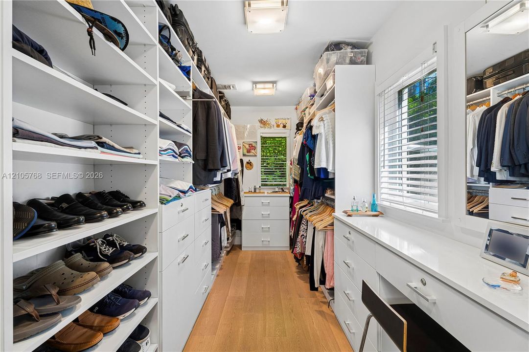 Custom walk-in closet featuring extensive built-in white shelving, integrated dresser drawers, wood-finish flooring, a dedicated vanity station, and large rectangular windows with horizontal blinds