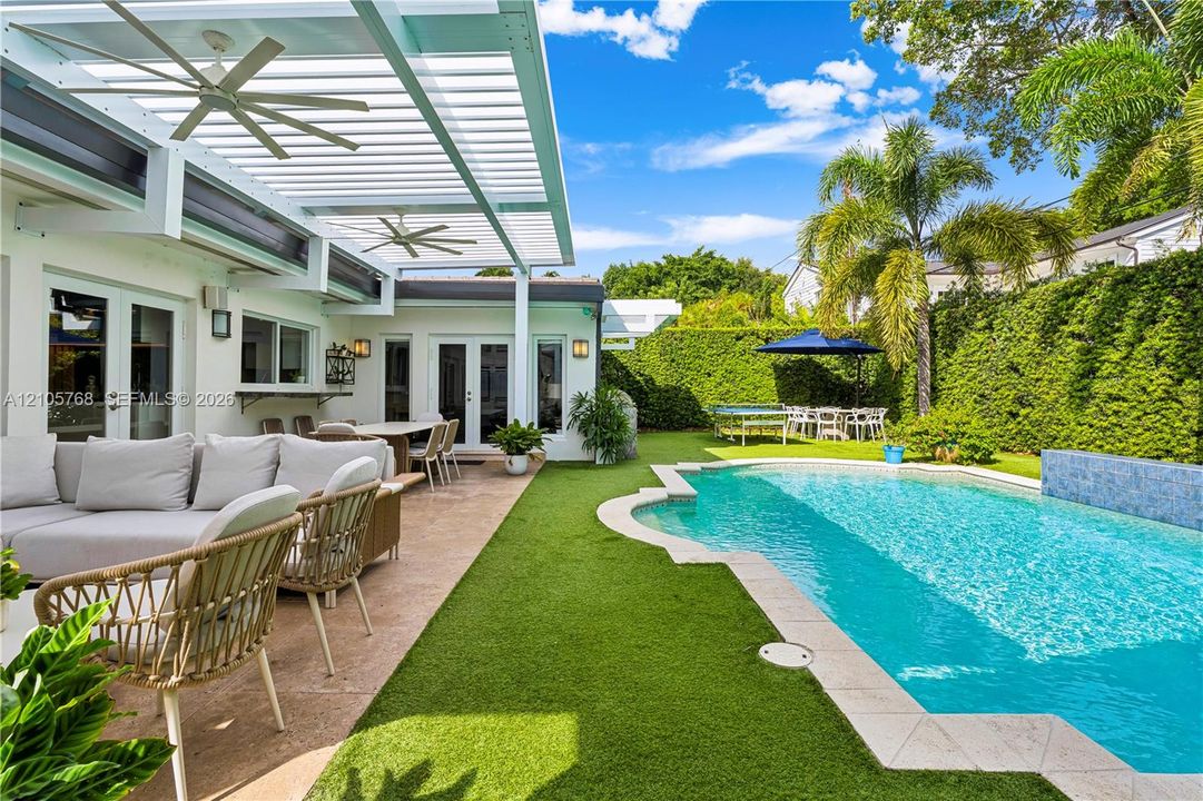 Outdoor living space featuring a white slatted pergola with ceiling fans, a custom-shaped swimming pool with a mosaic-tiled water feature, and artificial turf lawn bordered by a perimeter hedge