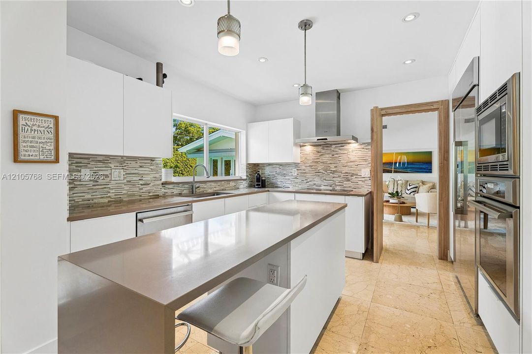 Kitchen featuring white flat-panel cabinetry, a solid-surface waterfall edge island, stacked-stone backsplash, built-in stainless steel wall oven and microwave, and recessed lighting