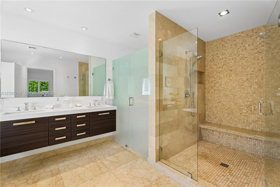 Floating dark wood-finish vanity with dual sinks, expansive wall-mounted mirror, walk-in glass-enclosed shower with mosaic tile wall, integrated stone bench, and recessed lighting