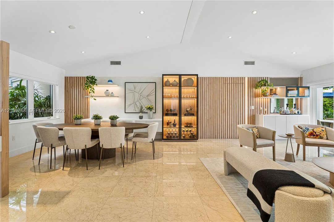 Open-concept living space featuring vaulted ceilings, polished stone flooring, custom vertical wood-slat wall paneling, built-in recessed shelving with integrated lighting, and a floor-to-ceiling glass-enclosed display cabinet
