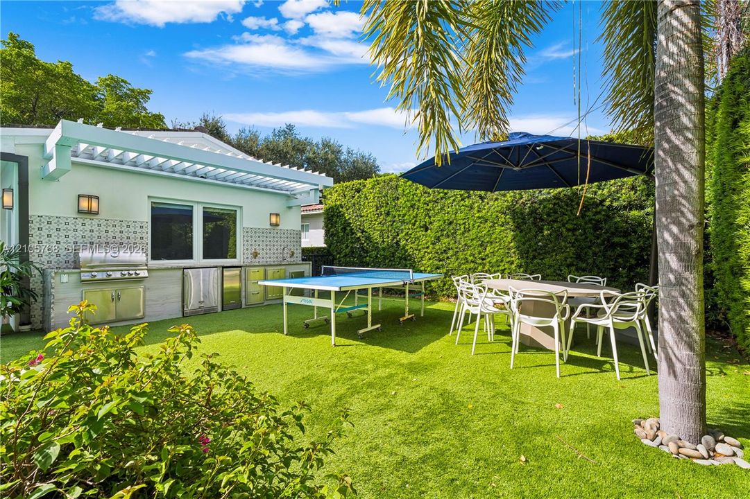 Outdoor kitchen featuring stainless steel appliances, patterned tile backsplash, custom cabinetry, a pergola-style roof structure, and artificial turf grounds