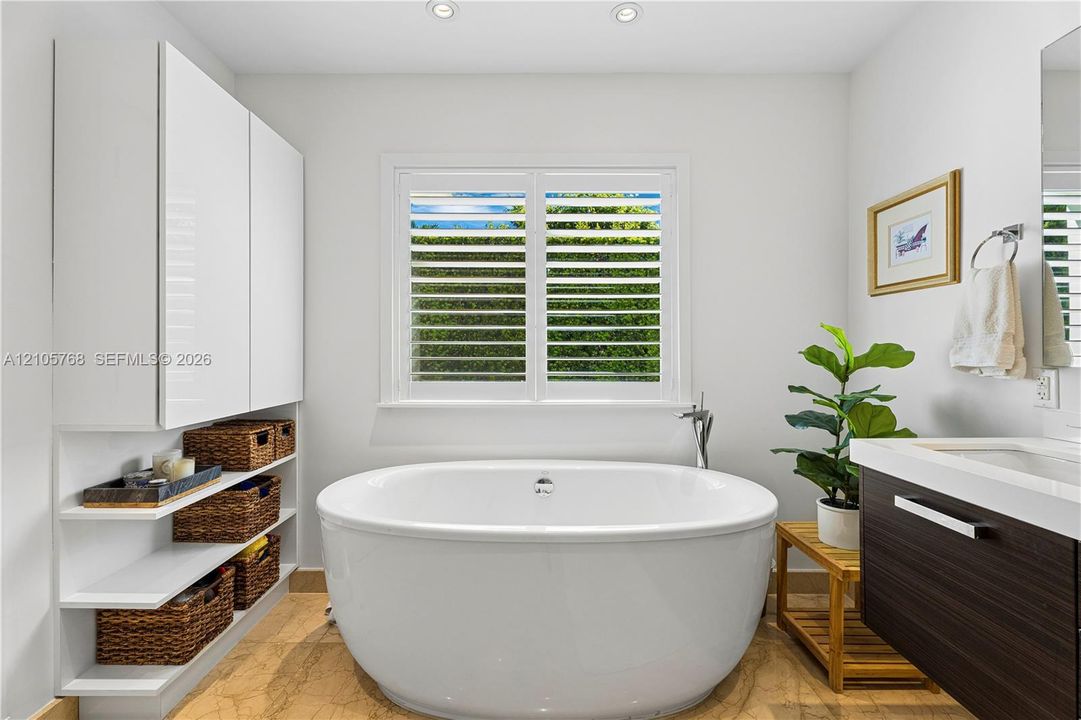 Freestanding soaking tub with a floor-mounted faucet, custom floor-to-ceiling cabinetry, open shelving, stone flooring, and a window fitted with interior plantation shutters