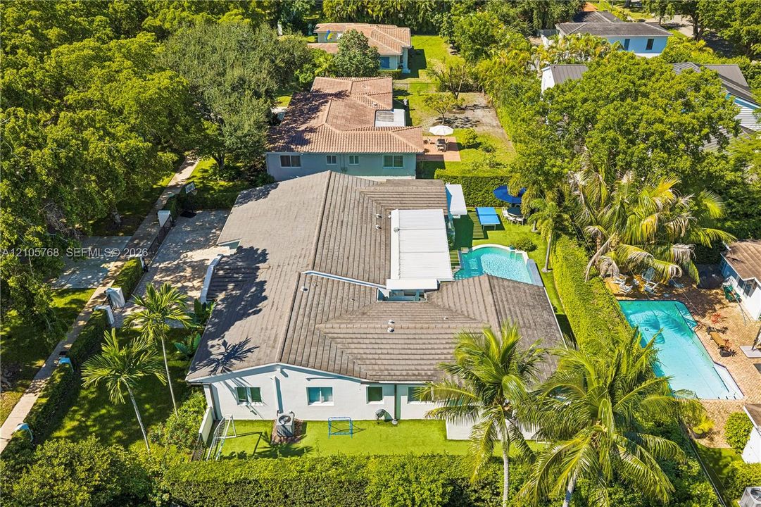 Aerial perspective of the residence featuring a multi-pitched shingled roof, a ground-level swimming pool, extensive landscaping, and concrete hardscaping