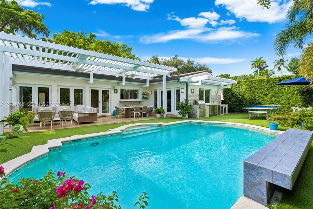 Outdoor living space featuring a white louvered pergola, an in-ground swimming pool with a built-in tiled bench, a stone-surfaced outdoor kitchen, and synthetic turf landscaping