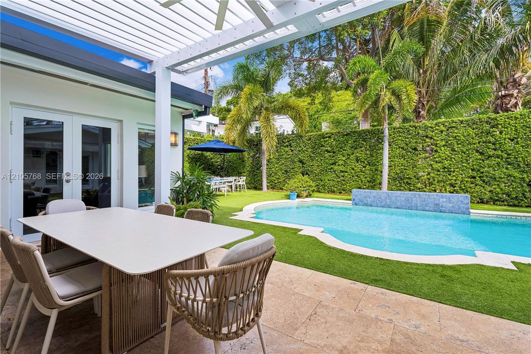 Covered patio with a white louvered pergola, stone-paver decking, private swimming pool featuring a tiled water feature, and maintained perimeter hedge landscaping