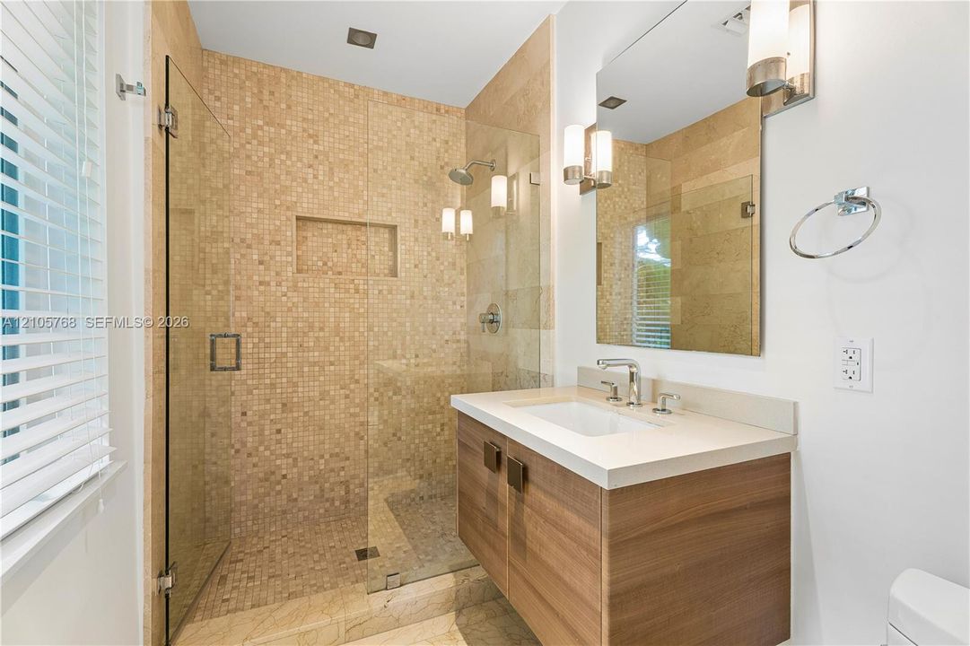 Bathroom featuring a wood-finish vanity with a stone countertop, a glass-enclosed shower with tiled walls and an integrated shower niche, and wall-mounted light fixtures
