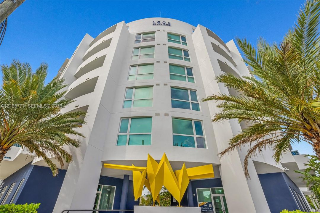 Mid-rise architectural exterior featuring a curved facade, private balconies, expansive windows, and a signature sculptural yellow entry feature