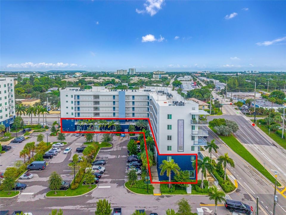 Multi-story residential building featuring a white and blue exterior, attached private balconies, surface-level parking, and mature palm landscaping