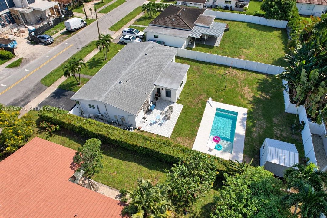 Aerial perspective of a property featuring an in-ground swimming pool, paved patio, expansive lawn, white vinyl perimeter fencing, and a detached storage structure