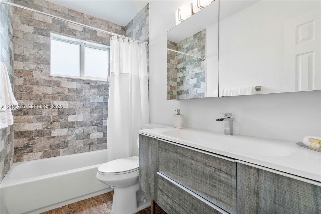 Textured stone tile wall encompassing the bathtub, wood-finish floating vanity, integrated rectangular sink, wall-mounted faucet, and mirrored medicine cabinet