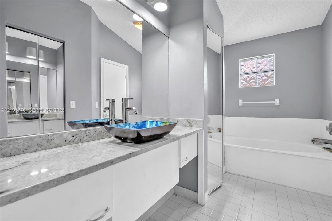 Stone-surface vanity with a decorative vessel sink, wall-spanning vanity mirror, floor-to-ceiling mirror panel, glass block window, and tile flooring