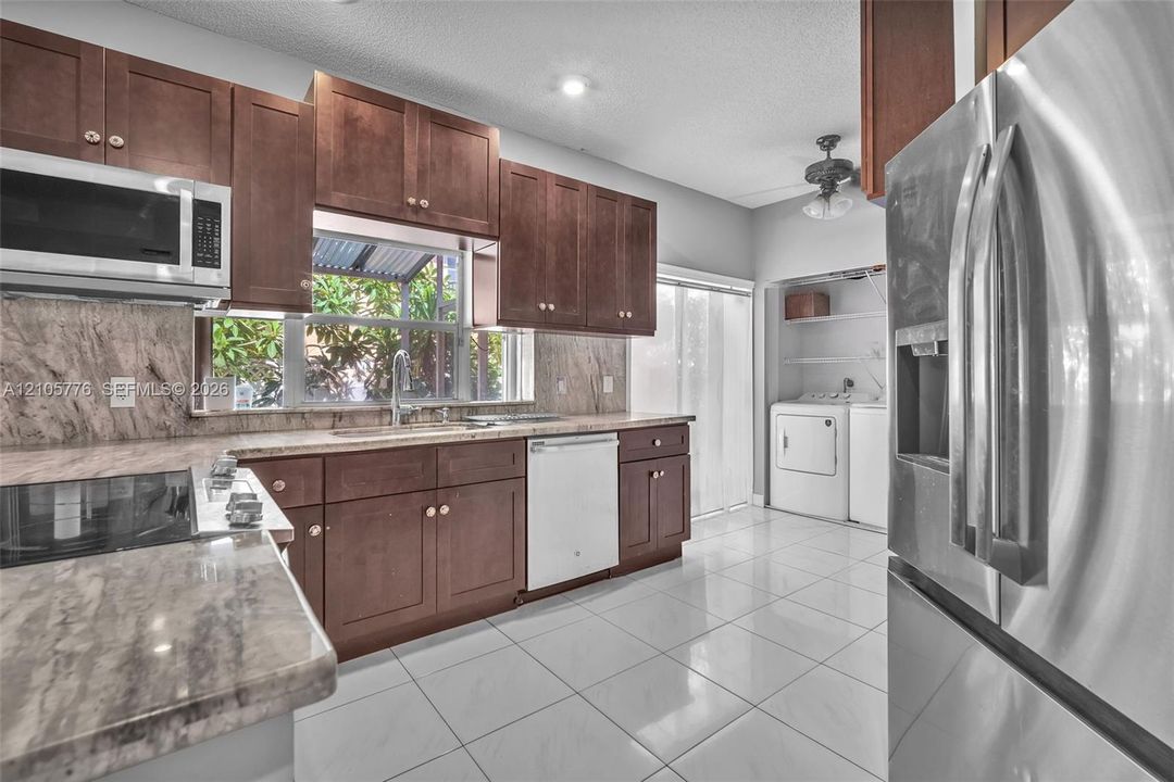 Kitchen featuring dark wood cabinetry, stone-patterned countertops, large-format tile flooring, stainless steel appliances, and a dedicated laundry area with built-in shelving