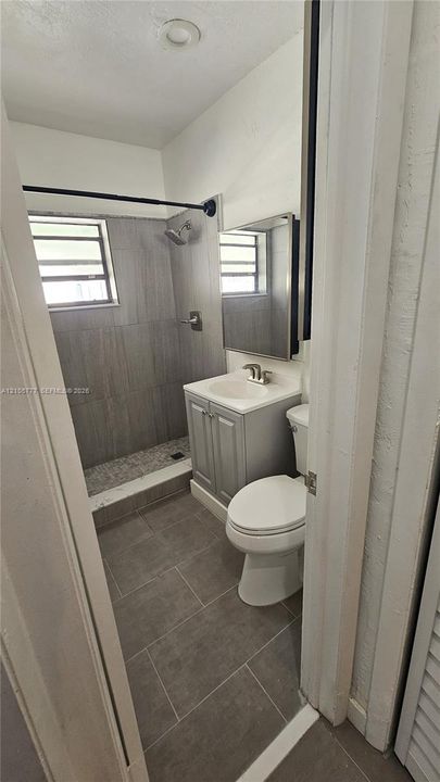 Bathroom featuring a walk-in shower with large-format grey tile, a single-door vanity with a composite countertop, an integrated rectangular sink, and neutral tile flooring
