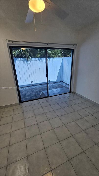 Ceramic tile flooring, ceiling fan with integrated light fixture, and black-framed sliding glass doors leading to a paved patio area with white privacy fencing