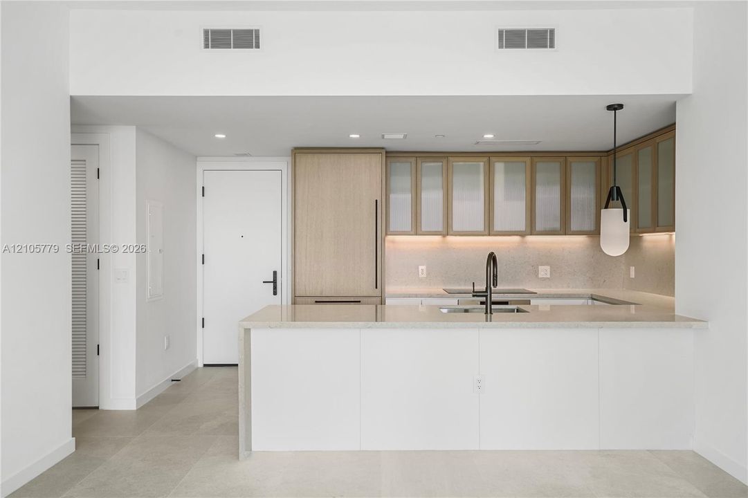 Kitchen featuring light wood-finish cabinetry with frosted glass inserts, a white quartz waterfall-edge island, integrated refrigerator, matte black gooseneck faucet, and under-cabinet lighting