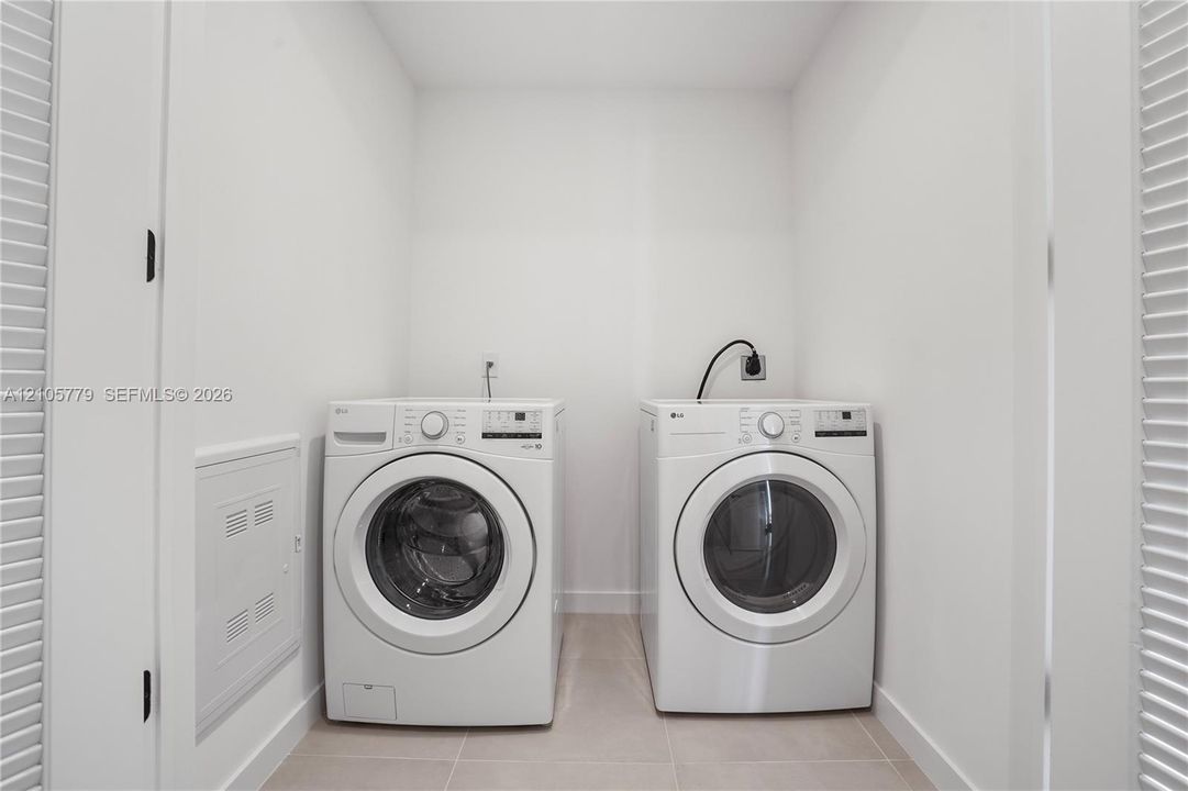 Laundry space featuring white front-loading appliances, neutral tile flooring, and louvered privacy doors