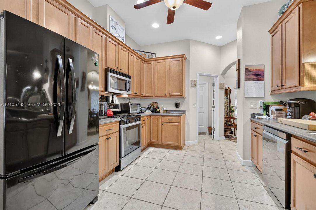 Kitchen featuring natural wood cabinetry, light-toned stone tile flooring, neutral countertops, stainless steel appliance suite, and a ceiling fan with integrated lighting
