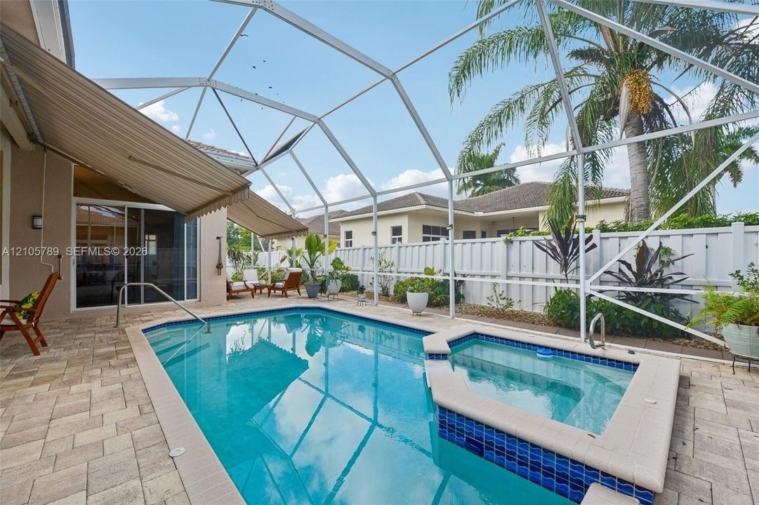 Screen-enclosed pool area featuring a spillover spa with blue tile accents, paver patio, retractable awning, and perimeter privacy fencing
