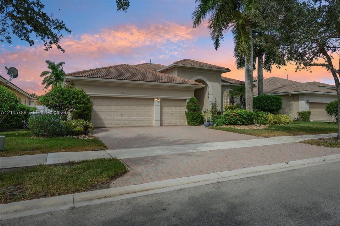 Stucco exterior with a tiled roof, arched entryway, and two-car garage featuring paver driveway and mature landscaping