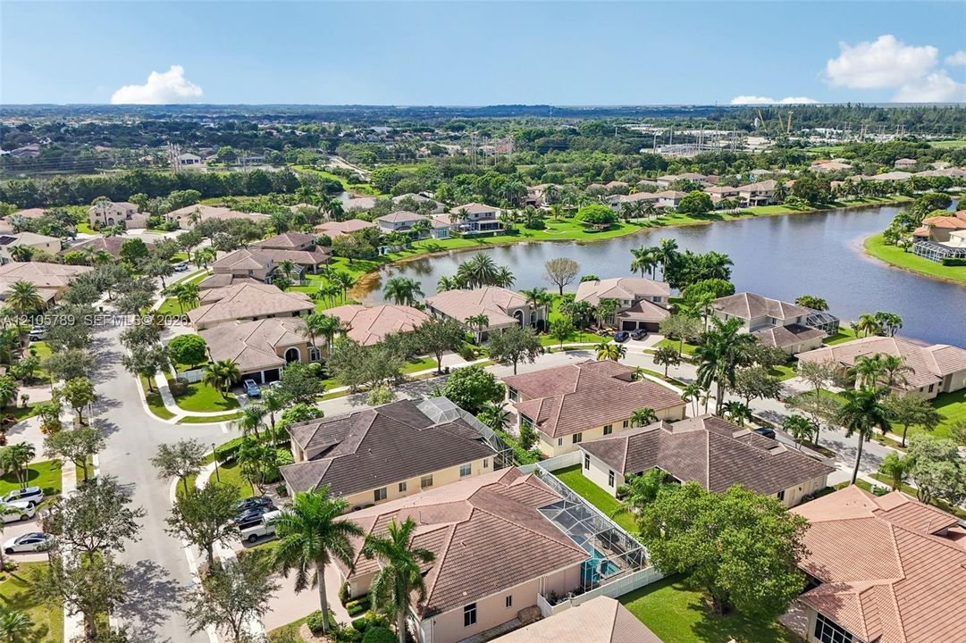 Aerial perspective of a residential community featuring expansive lake frontage, tile roofing, screened-in pool enclosures, and mature palm trees