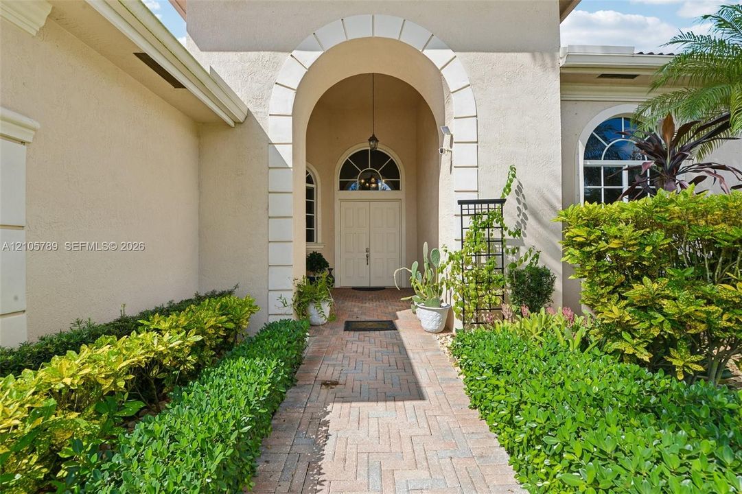 Stucco exterior entryway featuring a prominent arched portico with decorative quoins, double front doors with a semi-circular transom window, and a herringbone-patterned brick walkway