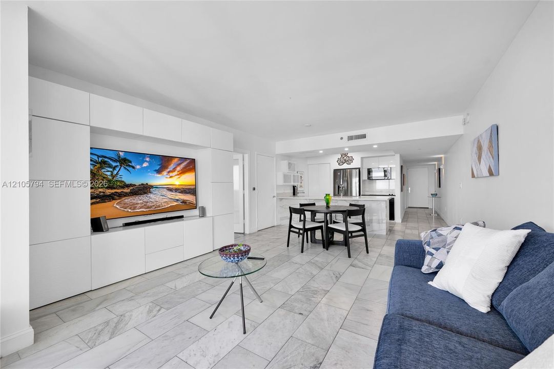 Living area featuring large-format stone tile flooring, custom white built-in media cabinetry, and an open kitchen with stone-topped island and white cabinetry