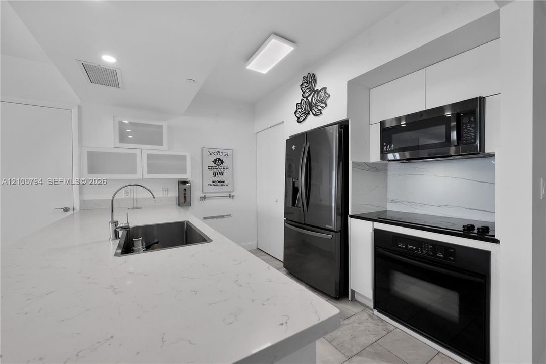 Kitchen featuring a central island with a stone-finish countertop and undermount sink, white cabinetry with frosted glass accents, stone-finish backsplash, built-in stainless steel microwave and oven, and dark-toned full-size refrigerator