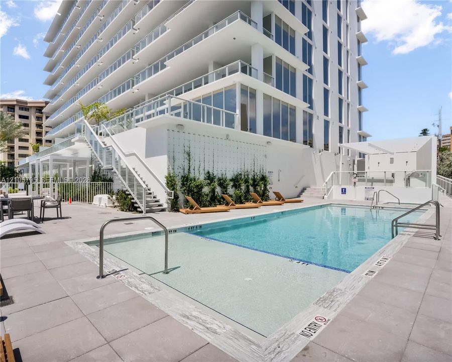 Swimming pool with a baja shelf, expansive concrete deck, metal railings, and exterior wall with integrated climbing vegetation