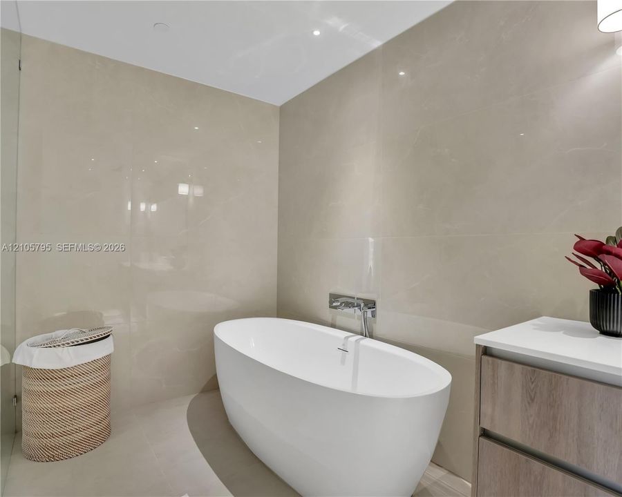 Freestanding white soaking tub with wall-mounted polished chrome faucet, floor-to-ceiling large-format tile wall cladding, and wood-finish vanity cabinet with a white countertop