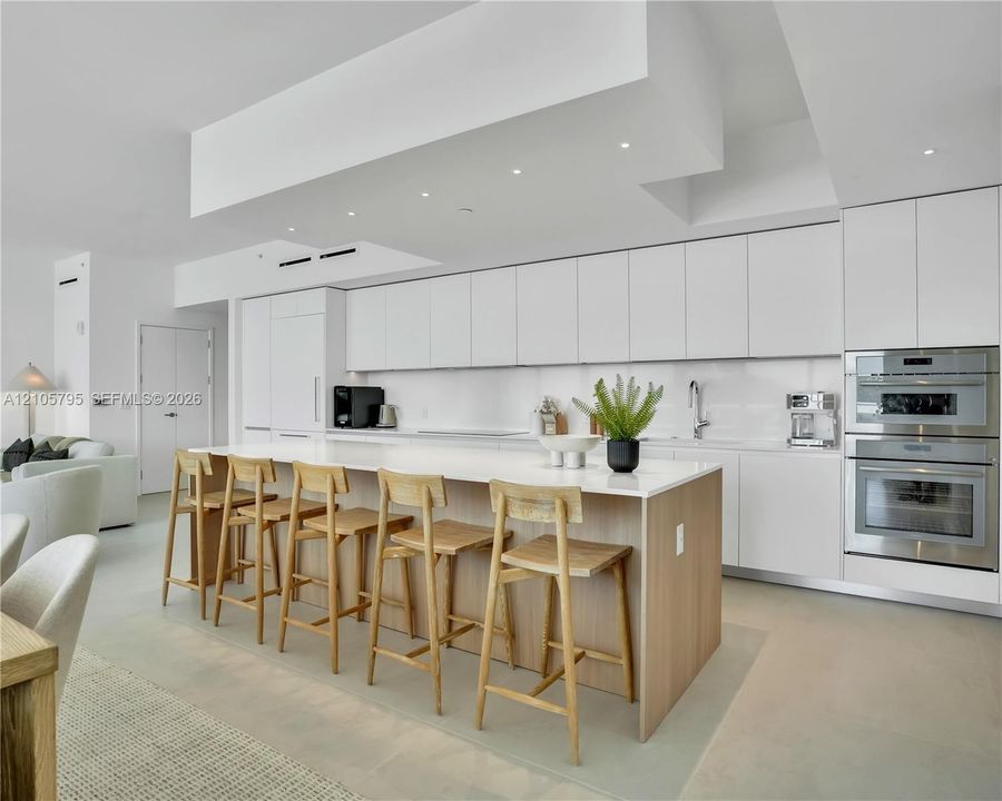 Modern kitchen featuring seamless flat-panel white cabinetry, a waterfall-edge central island with wood-finish base, integrated stainless steel double wall ovens, recessed ceiling lighting, and smooth-finish flooring