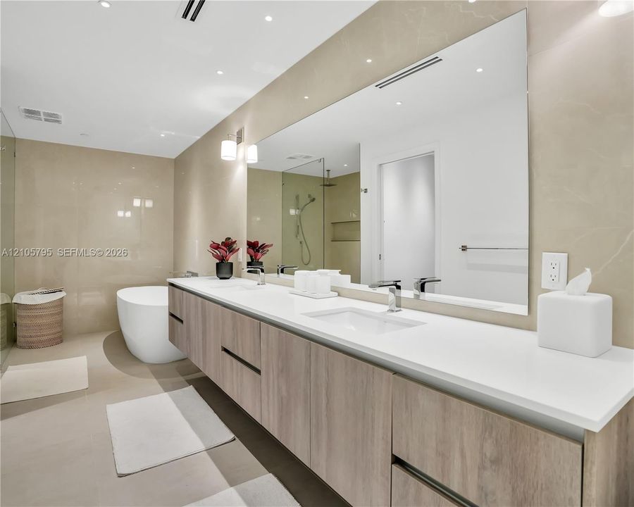 Floating dual-sink vanity featuring wood-finish cabinetry, solid surface countertops, polished chrome fixtures, and a large frameless wall-to-wall mirror, complemented by a freestanding soaking tub and full-height tiled wall surround