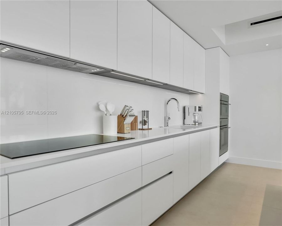 Minimalist kitchen featuring matte white flat-panel cabinetry, smooth white countertops, integrated cooktop, chrome gooseneck faucet, and stainless steel built-in wall oven