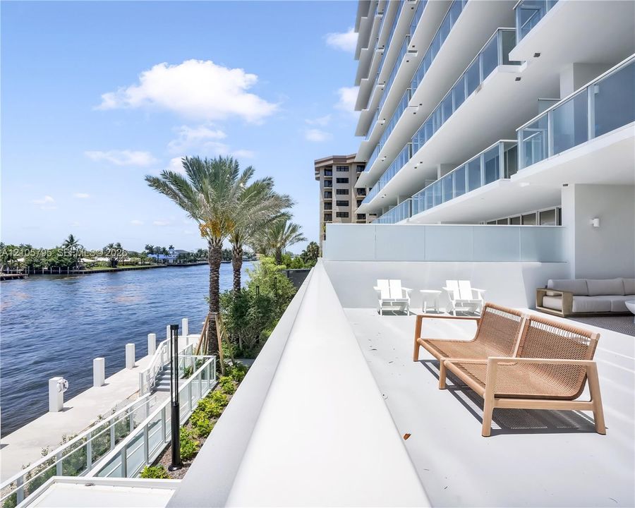 Expansive private balcony featuring glass panel railings, concrete flooring, and direct water frontage overlooking the waterway, complemented by a stone-paved bulkhead and integrated lighting