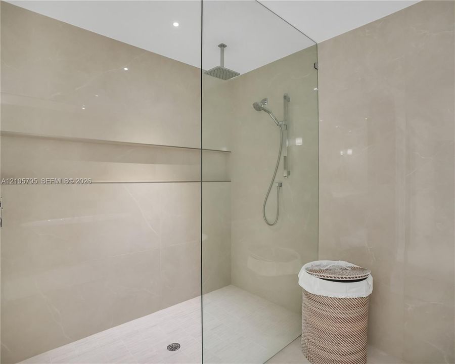 Walk-in shower featuring large-format stone-finish tile surround, integrated recessed shelving, a frameless glass enclosure, a ceiling-mounted rainfall showerhead, and a hand-held shower wand