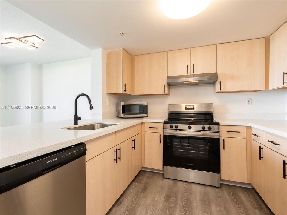 Kitchen featuring light wood-finish cabinetry, white countertops, stainless steel appliances, matte black hardware, and wood-finish flooring