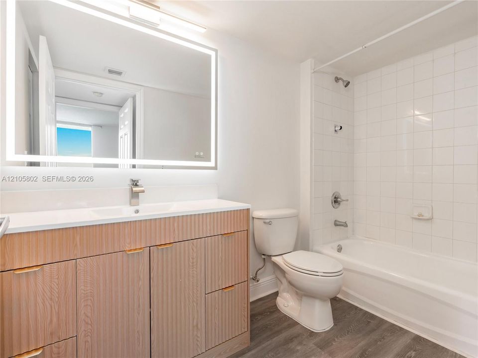 Bathroom featuring a light wood-finish vanity with integrated sink, rectangular LED-backlit mirror, wood-finish flooring, and white wall-tile surround at the shower-tub combination