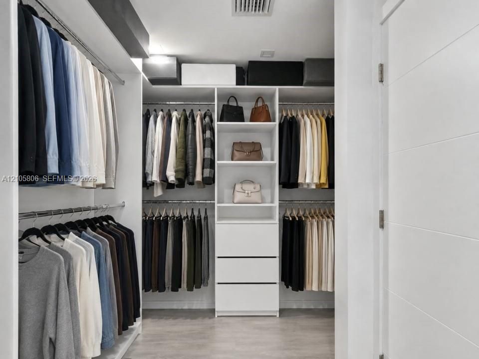 Custom walk-in closet featuring white built-in shelving, a central cabinetry unit with three drawers, dual-tier metal hanging rods, and wood-finish flooring