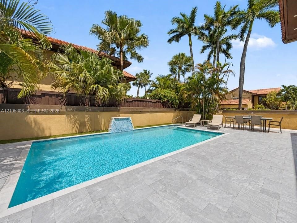 Rectangular swimming pool featuring a decorative tiled water feature, surrounded by expansive stone paver decking and perimeter privacy walls shaded by mature palm trees