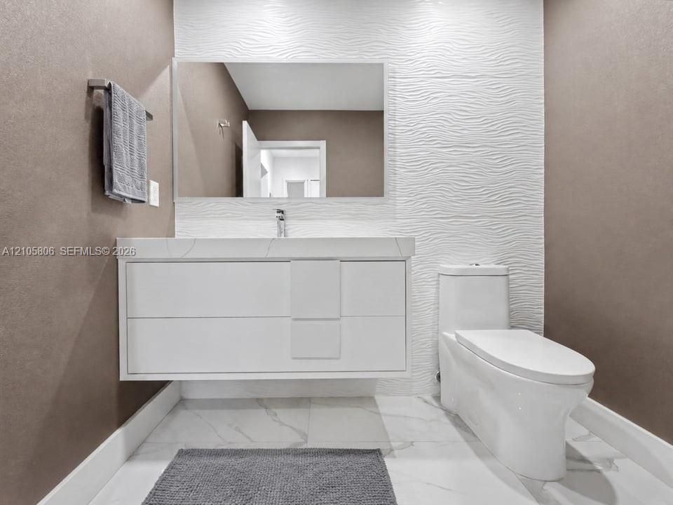 Floating vanity with integrated sink, stone-surface countertop, textured white feature wall, large-format tiled flooring, and dual-flush toilet