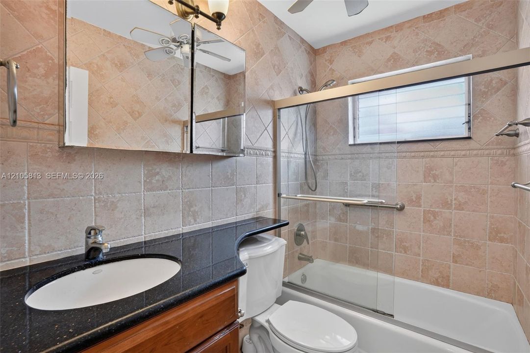 Bathroom featuring stone-finish wall tile, dark stone countertop with integrated oval sink, wood-finish vanity cabinetry, and a bathtub with a glass sliding door enclosure