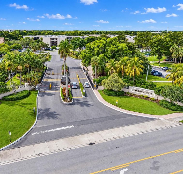 Gated entrance featuring a security checkpoint, lush landscaping with mature palms, manicured lawns, and paved thoroughfares