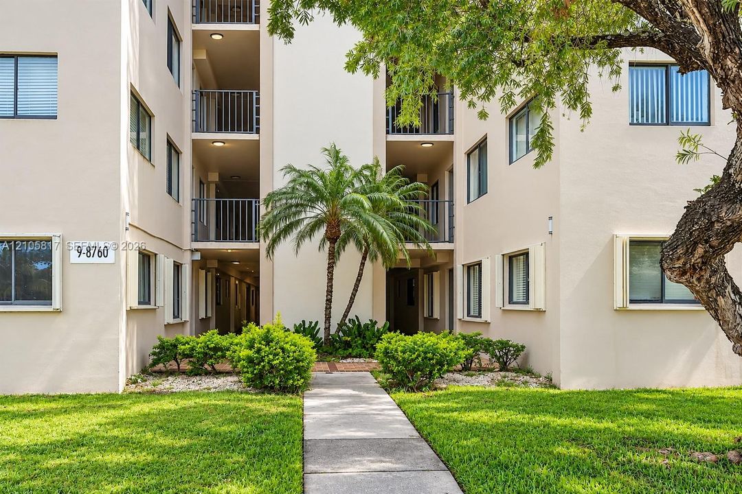 Multi-story stucco exterior featuring stacked balconies with metal railings, a central paved walkway, mature palm trees, and manicured landscaping