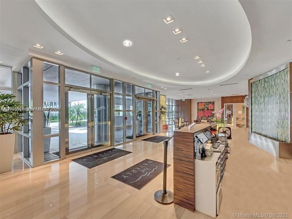 Lobby featuring high-gloss tile flooring, an integrated tiered reception desk with wood-grain veneer, a curved recessed ceiling with perimeter cove lighting, and a textured decorative water feature