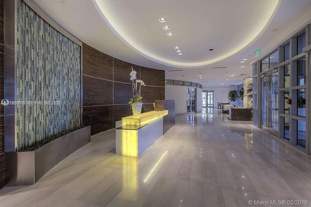Lobby featuring a vertical glass tile water feature, curvilinear wood-finish wall paneling, polished stone flooring, recessed cove lighting, and a built-in illuminated stone-topped reception desk