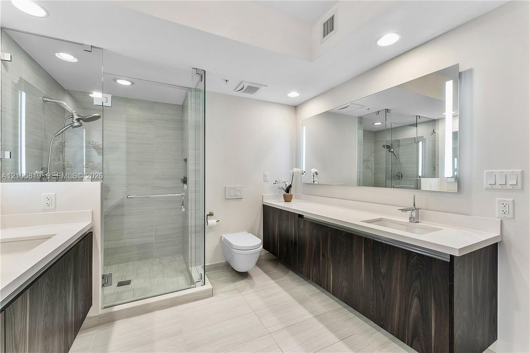 Floating dark wood-finish dual vanity with solid surface countertops, frameless glass-enclosed shower, wall-mounted toilet, and large integrated vanity mirror with LED lighting