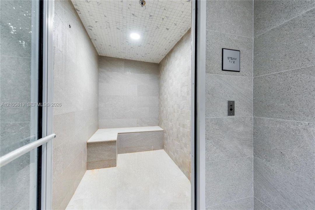 Steam room featuring large-format stone wall tile, mosaic ceiling tile, built-in stone bench, and glass-entry door with integrated control panel