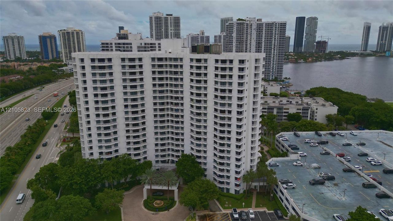 High-rise residential building featuring stacked private balconies, a covered porte-cochère entrance, paved circular driveway, and an attached multi-level parking structure