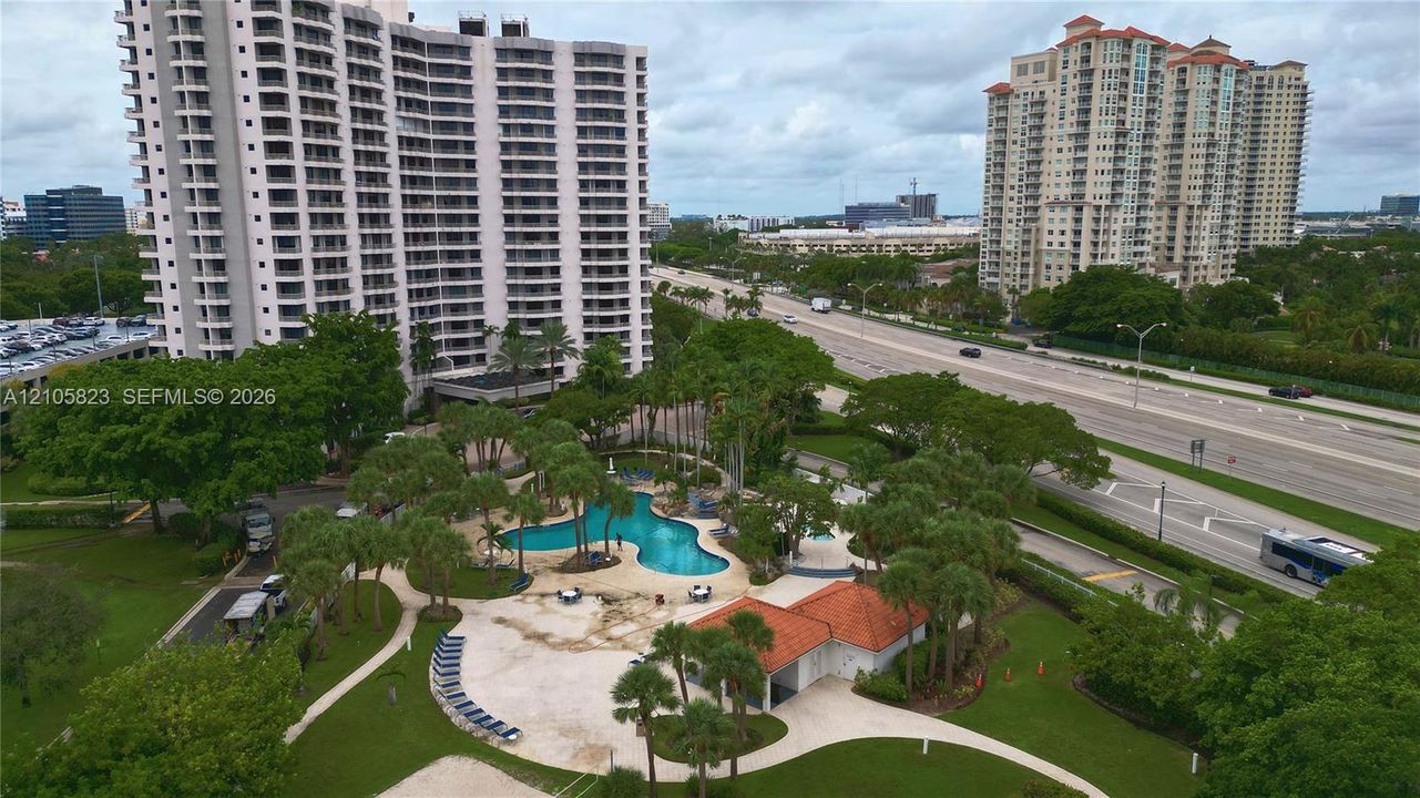 Outdoor common area featuring a freeform swimming pool, paved sundeck, terracotta-tiled pool house, manicured green space, and mature palm trees