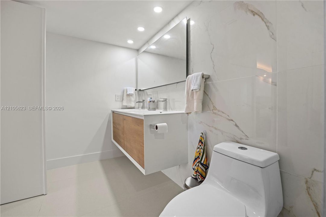Floating vanity with wood-finish cabinetry, rectangular vessel sink, large wall-mounted mirror, recessed ceiling lighting, and floor-to-ceiling porcelain wall tile